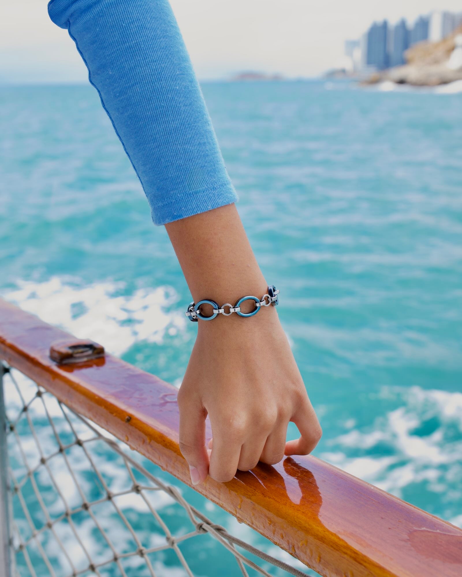 Women's wrist wearing Meridian Chain Bracelet Glacier Blue, knuckles resting on wooden boat railing, sea backdrop