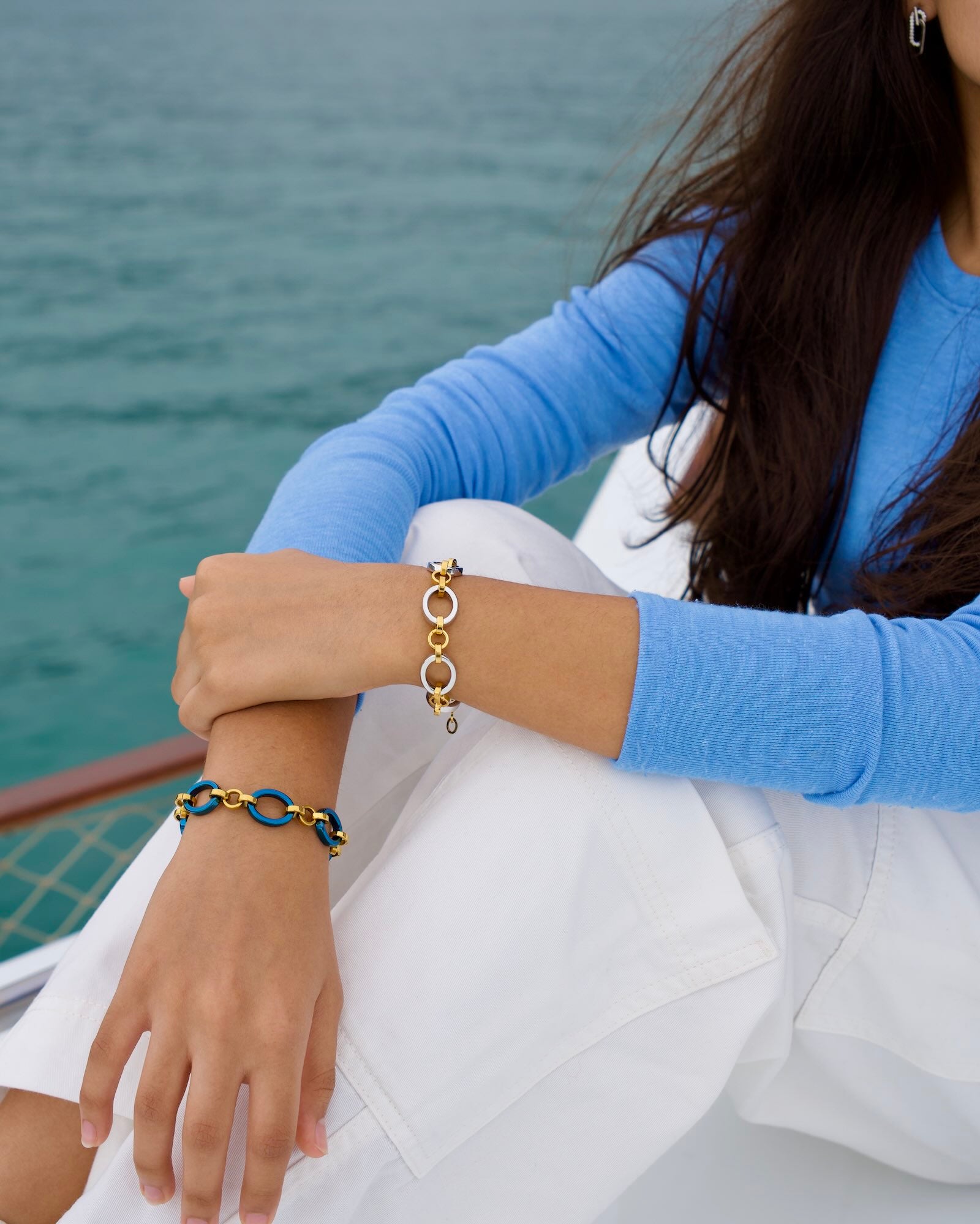 Women's wrists wearing Meridian Chain Bracelet in Midnight Gold and Twilight, seated on boat with sea backdrop