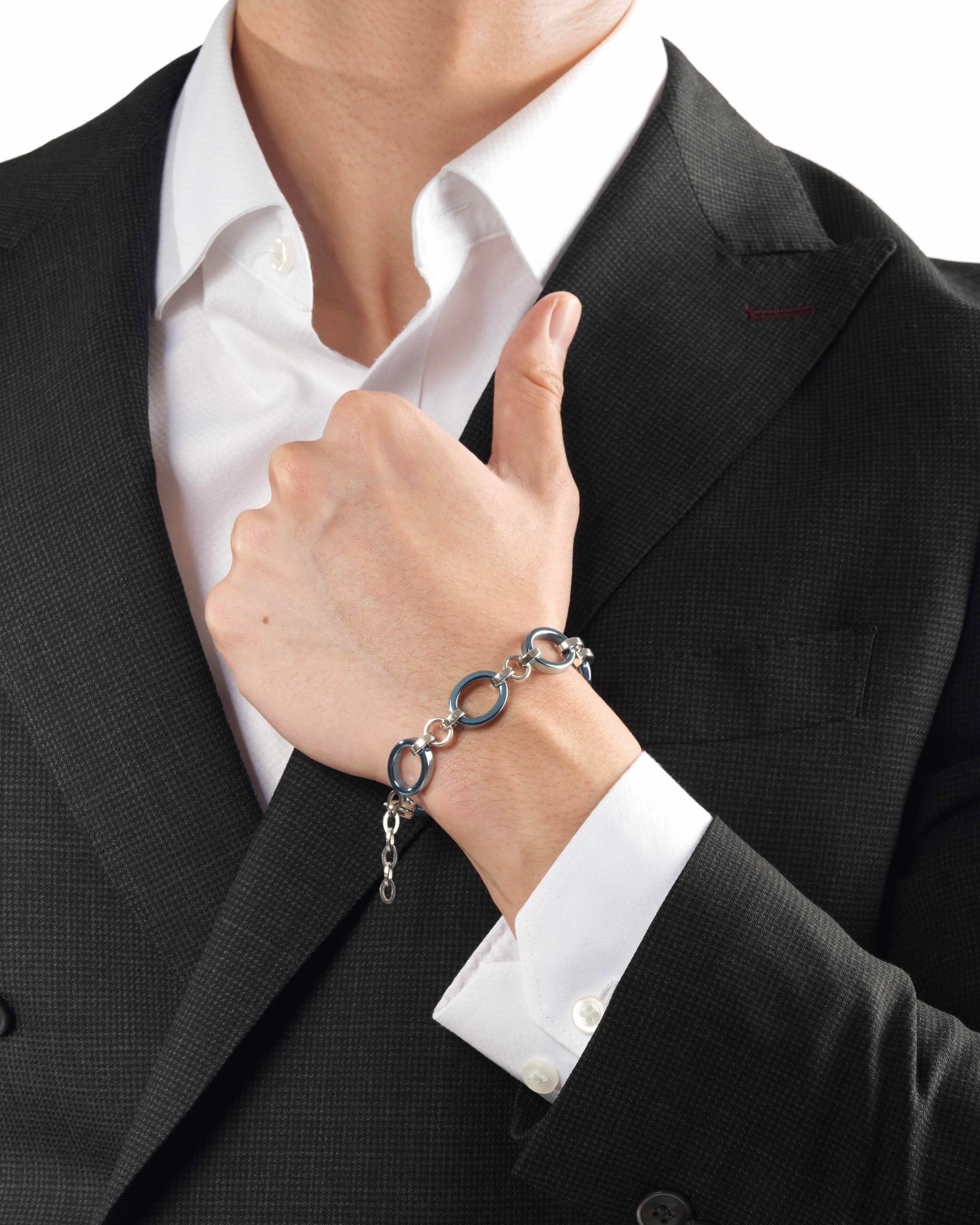 Man wearing a black suit with a white shirt and a ice blue and white gold chain bracelet on a plain background