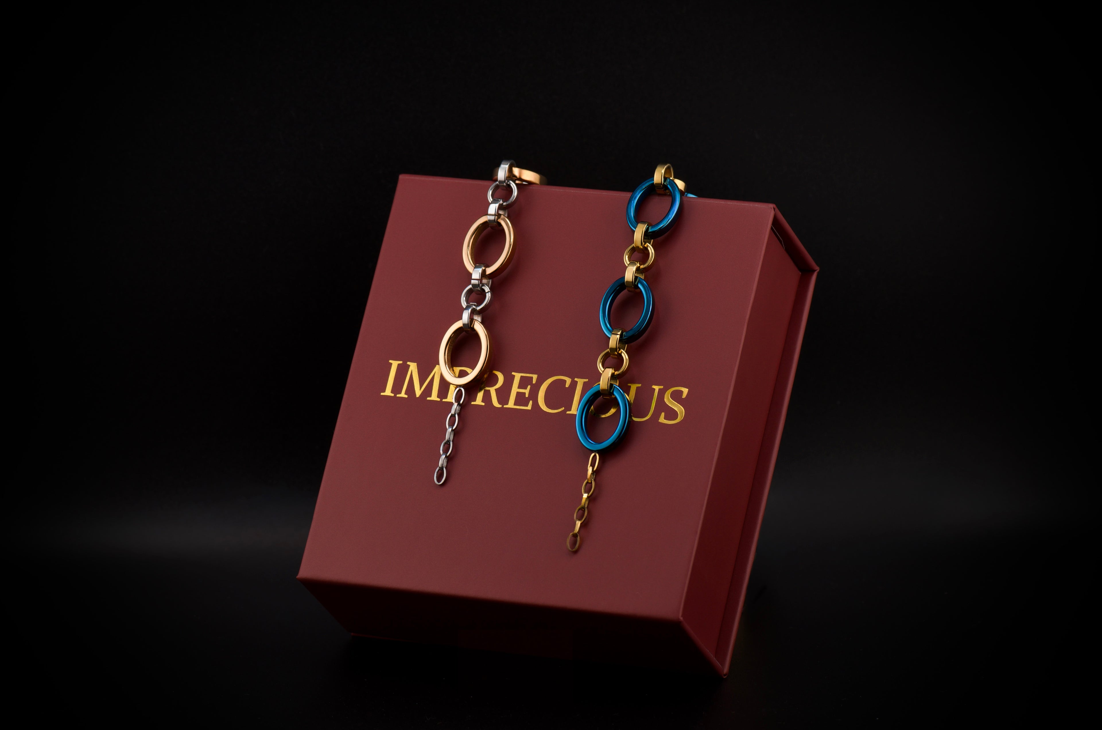 Pair of bracelets hanging on a maroon box with 'IMPRECIOUS' branding against a dark background